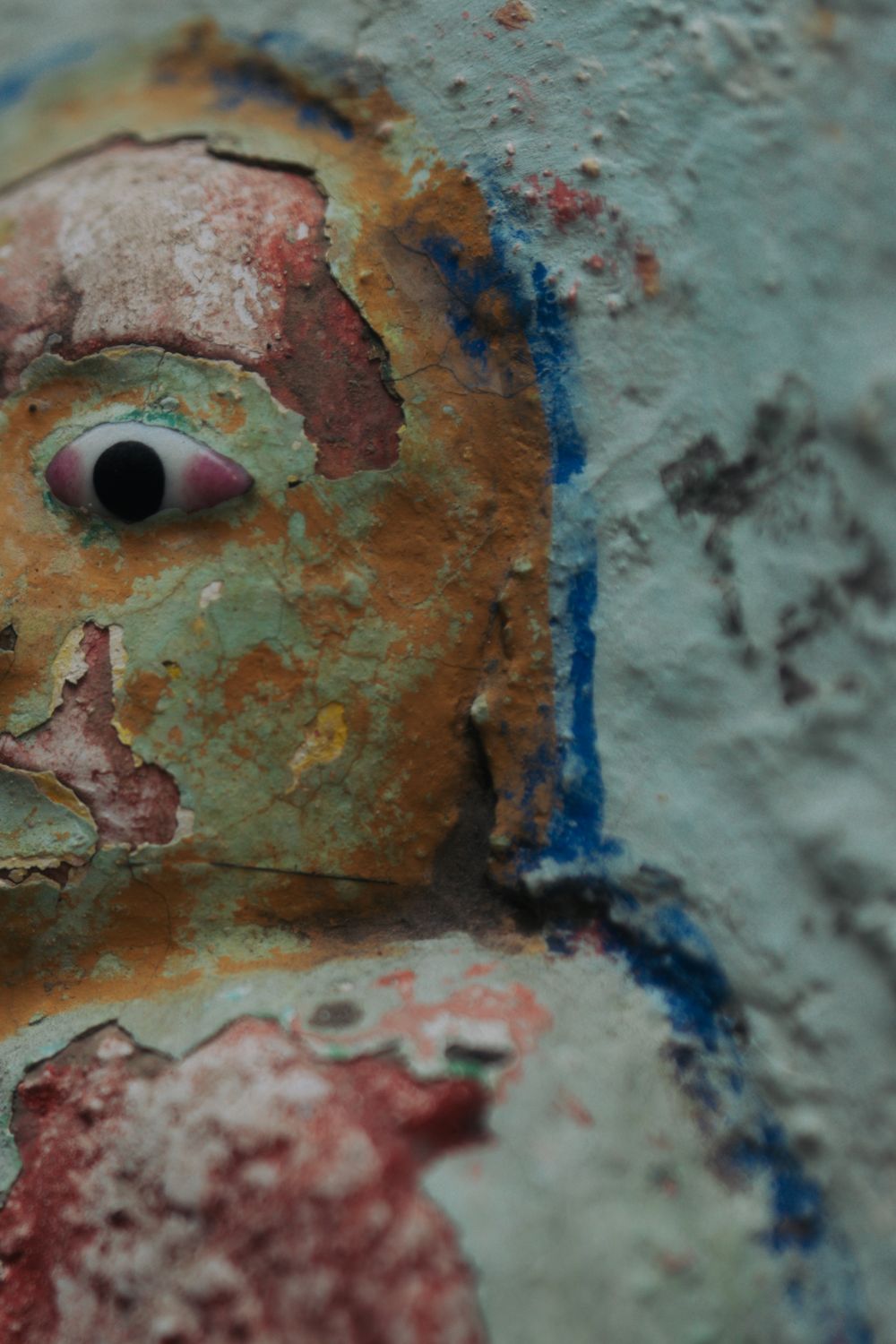 Close-up of weathered wall art showing distressed painted face with prominent eye.