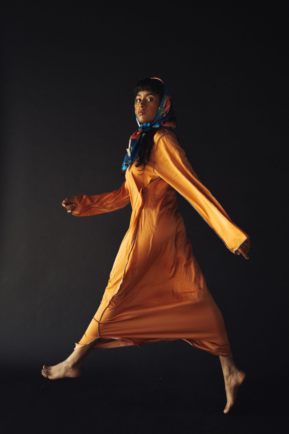 Figure in flowing orange dress and blue scarf leaping against dark background.