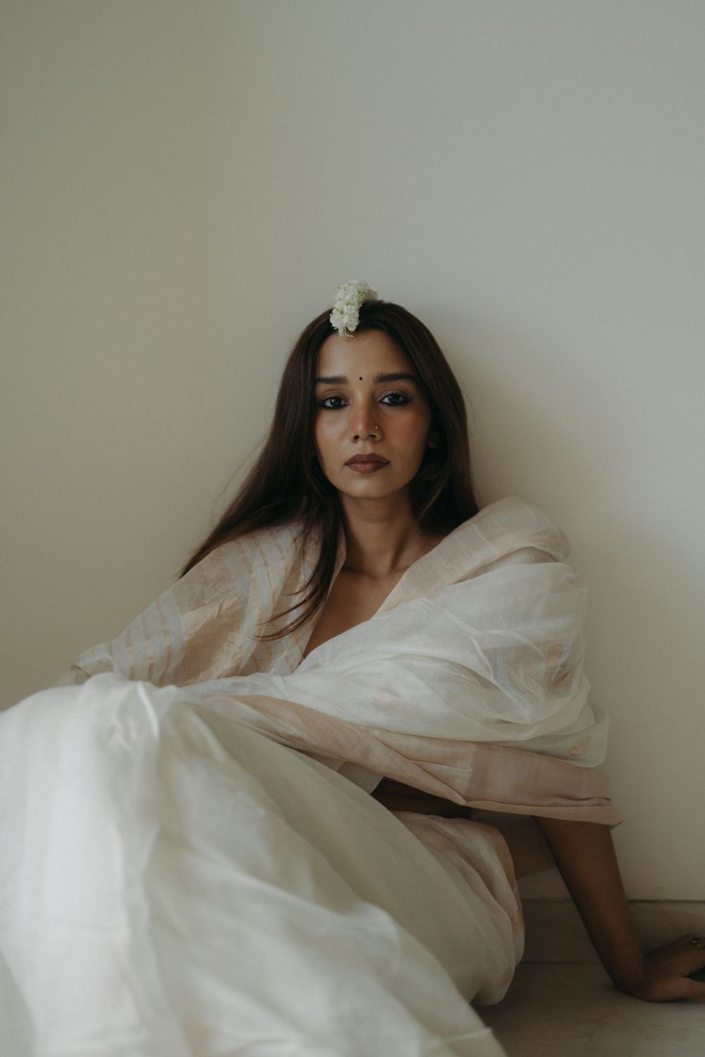 Ethereal portrait of woman in flowing white dress and delicate hair accessory against neutral wall.