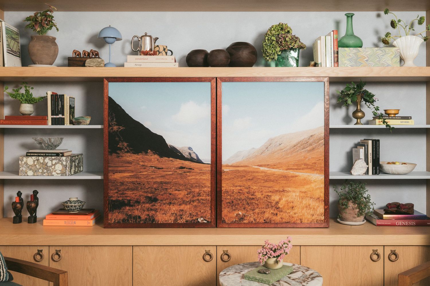Modern wooden shelving unit decorated with plants and landscape artwork displaying desert mountains against a white wall.