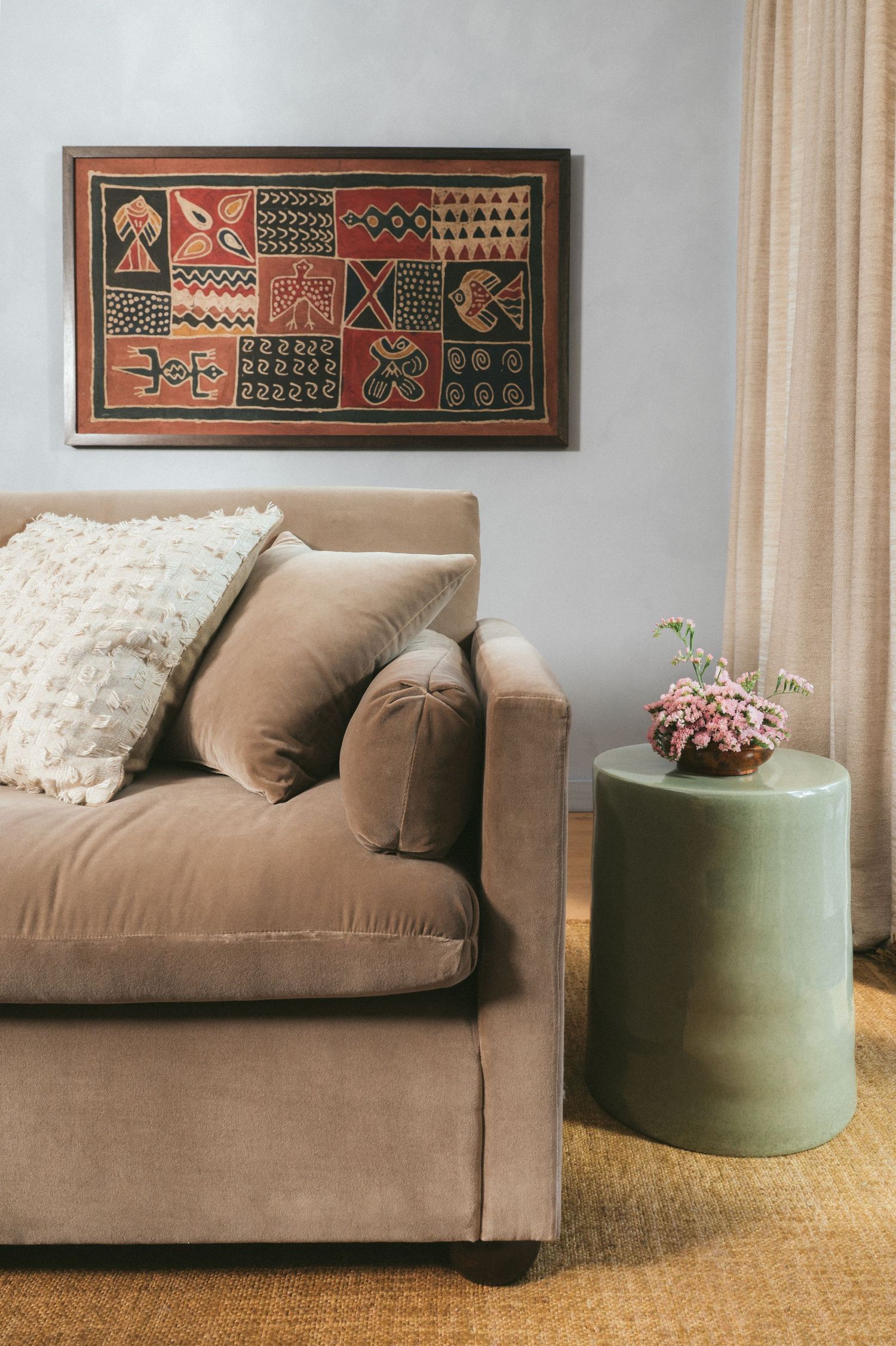 Contemporary living room with beige velvet sofa, green ottoman, and tribal wall art against gray walls.