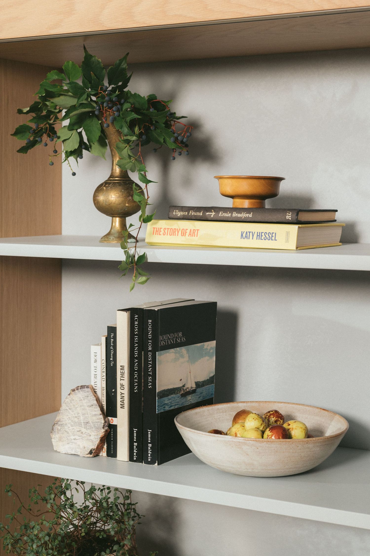 Styled bookshelf with decorative brass vase, stacked books, and stone bowl of fresh fruit on white shelves.