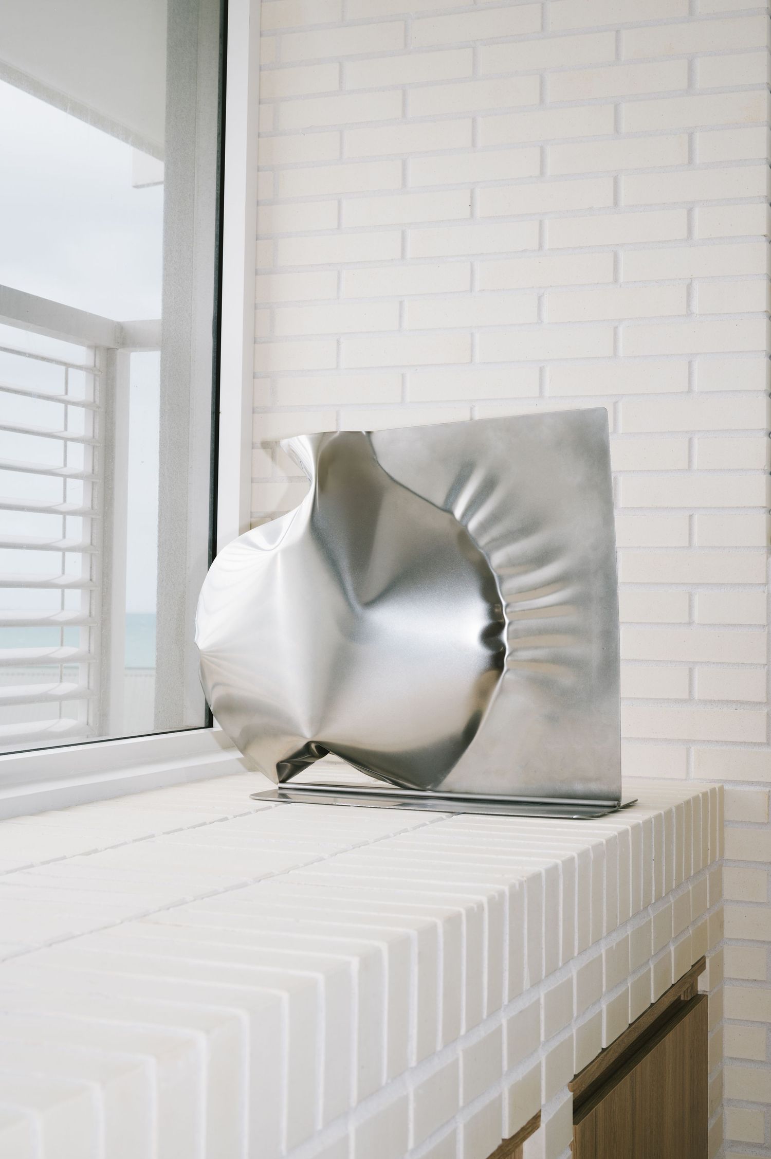 Abstract metallic sculpture with curved surfaces displayed on white tiled windowsill.