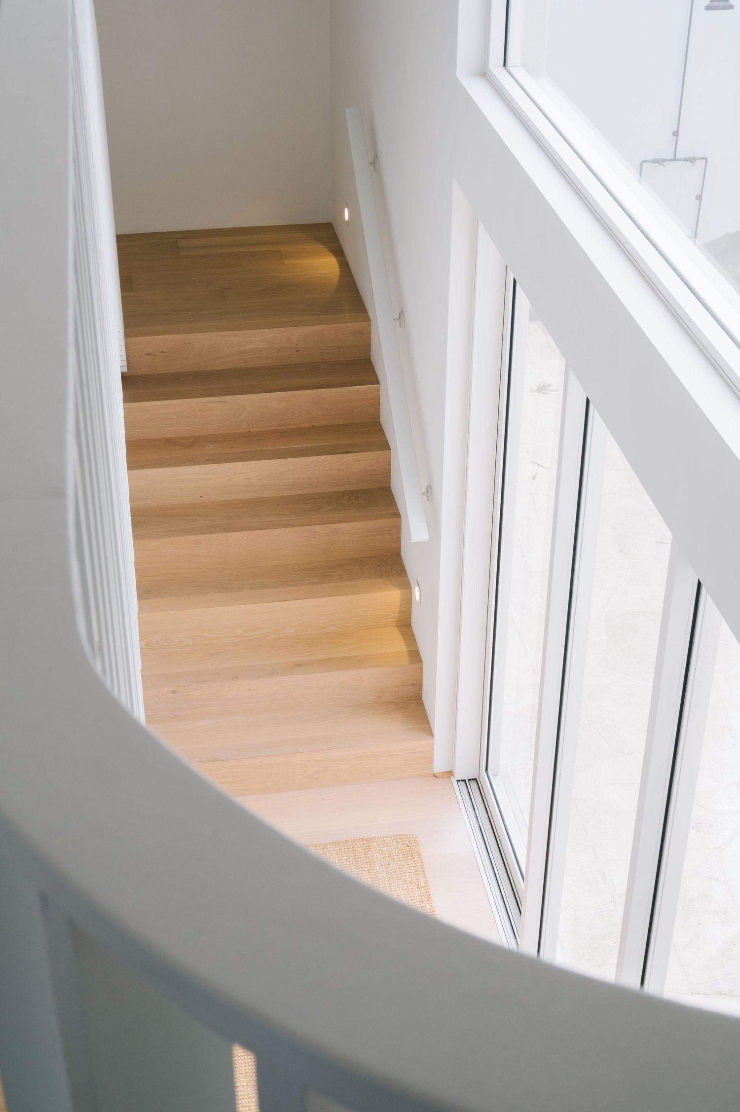 Light-filled stairwell with natural wood steps and white walls featuring large windows.