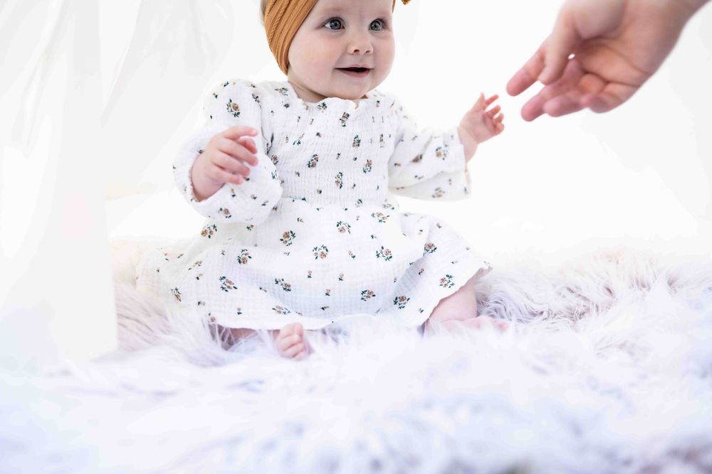 Cute baby in white floral romper with orange bow headband reaching toward an outstretched hand on fur.