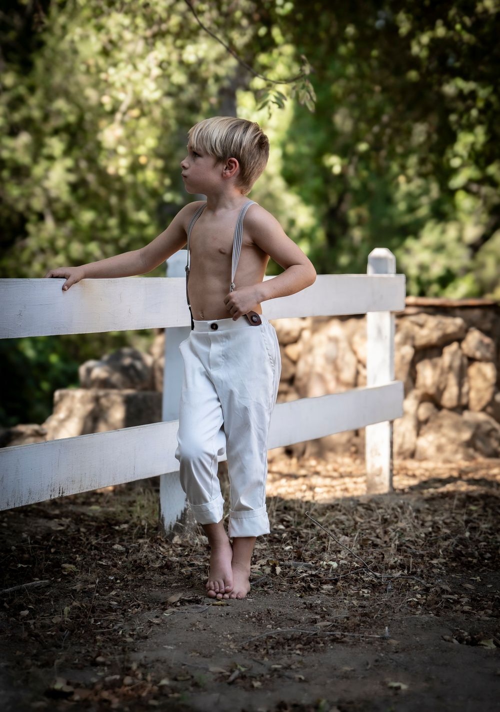 Shirtless young boy in linen pants and suspenders standing barefoot by a white wooden fence outdoors.