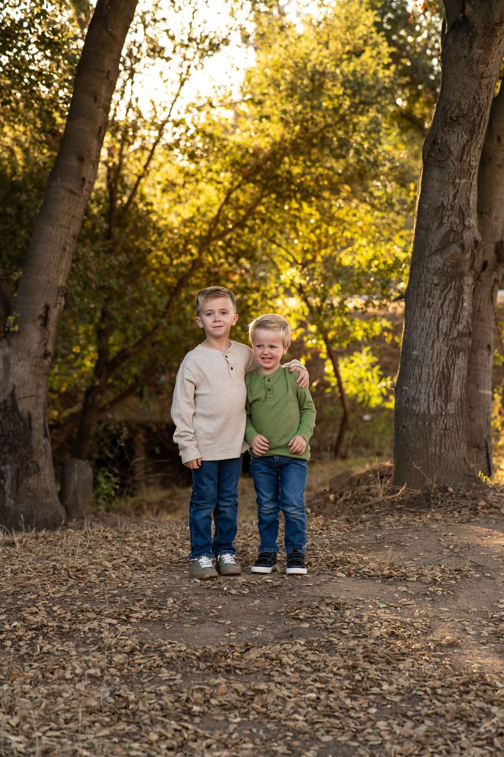 Two young brothers pose together on a forest path surrounded by autumn trees in golden light.