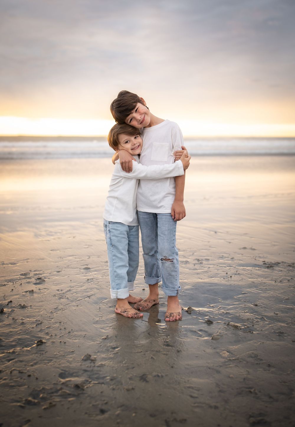 Two children hugging on a beach at sunset, wearing white tops and jeans, standing on wet sand. Solstice & Willow photography