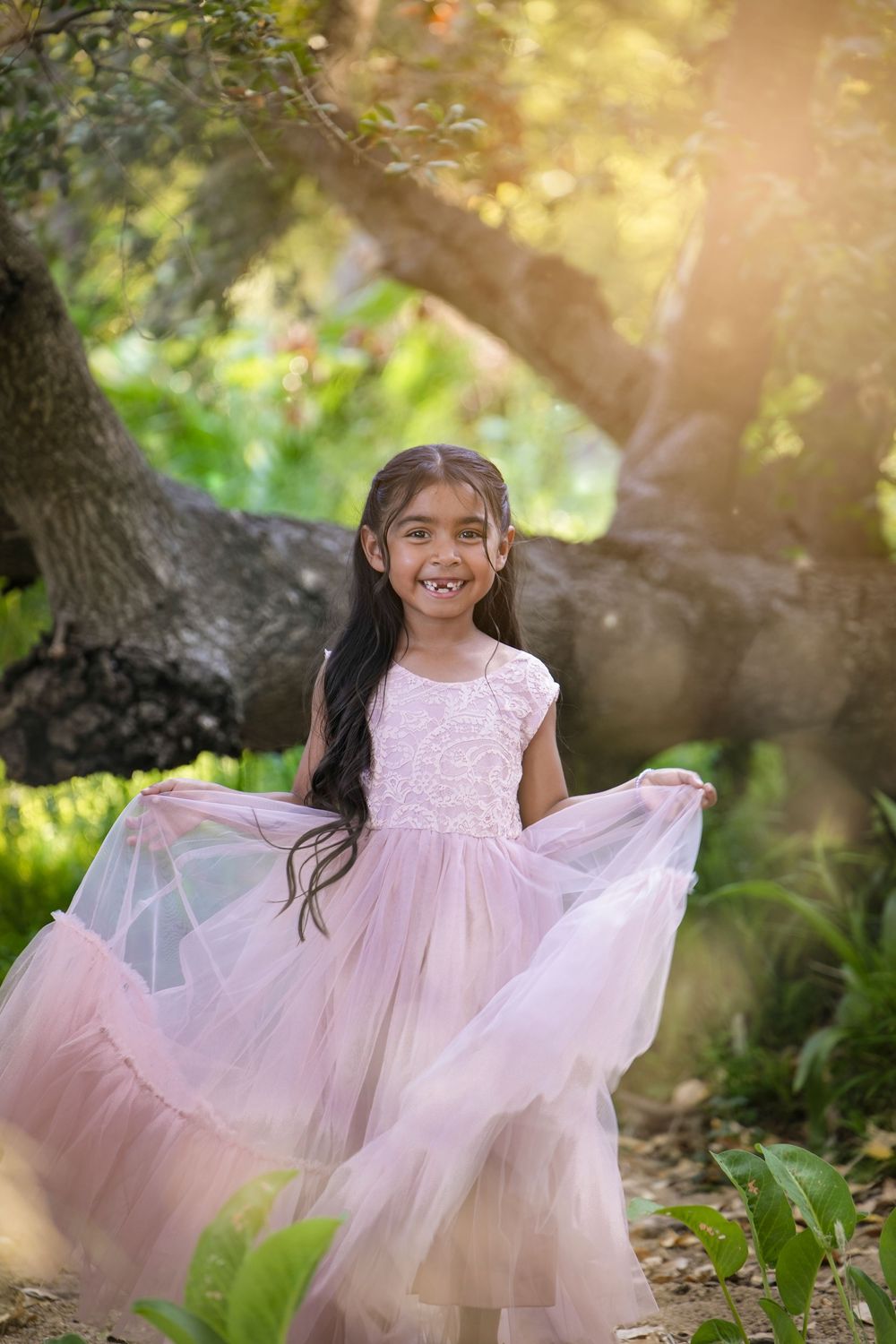 Young girl in pink tulle dress twirling outdoors in a sunlit garden, smiling joyfully among lush green trees. San Diego county