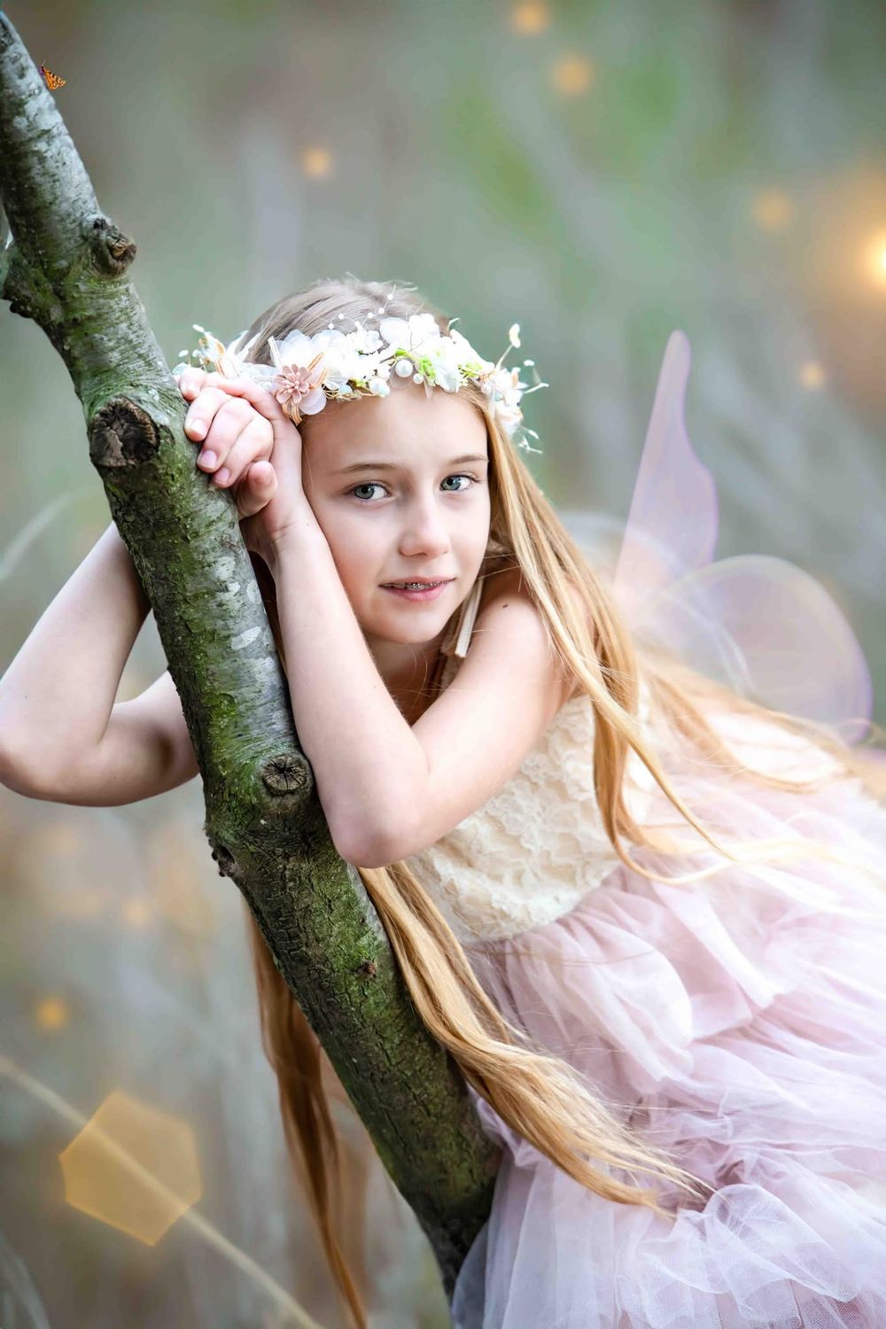 A young girl in a pink tutu dress and flower crown leans against a tree branch, dressed as a fairy with wings.