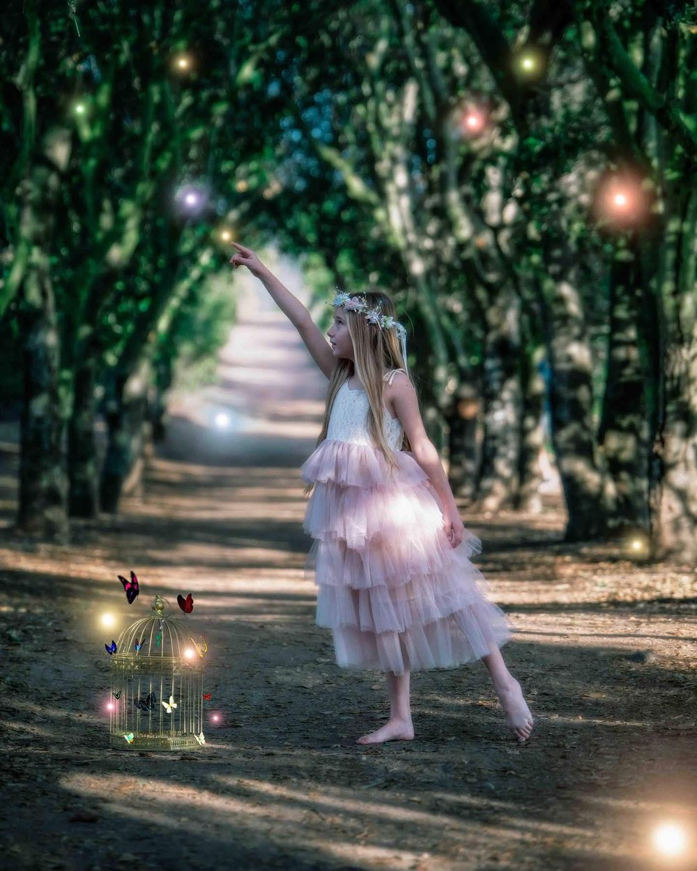 A young girl in a pink tutu dress reaches toward glowing fairy lights in an enchanted forest path with a decorative birdcage.