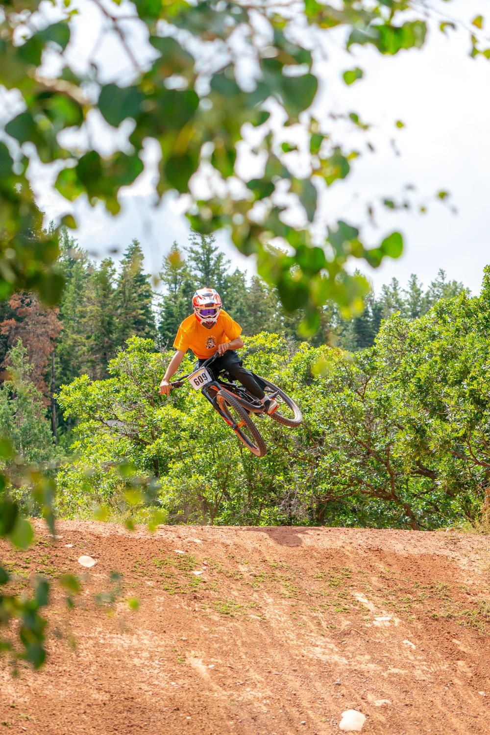 A cyclist wearing an orange shirt and helmet is mid-air while performing a jump on a dirt path.