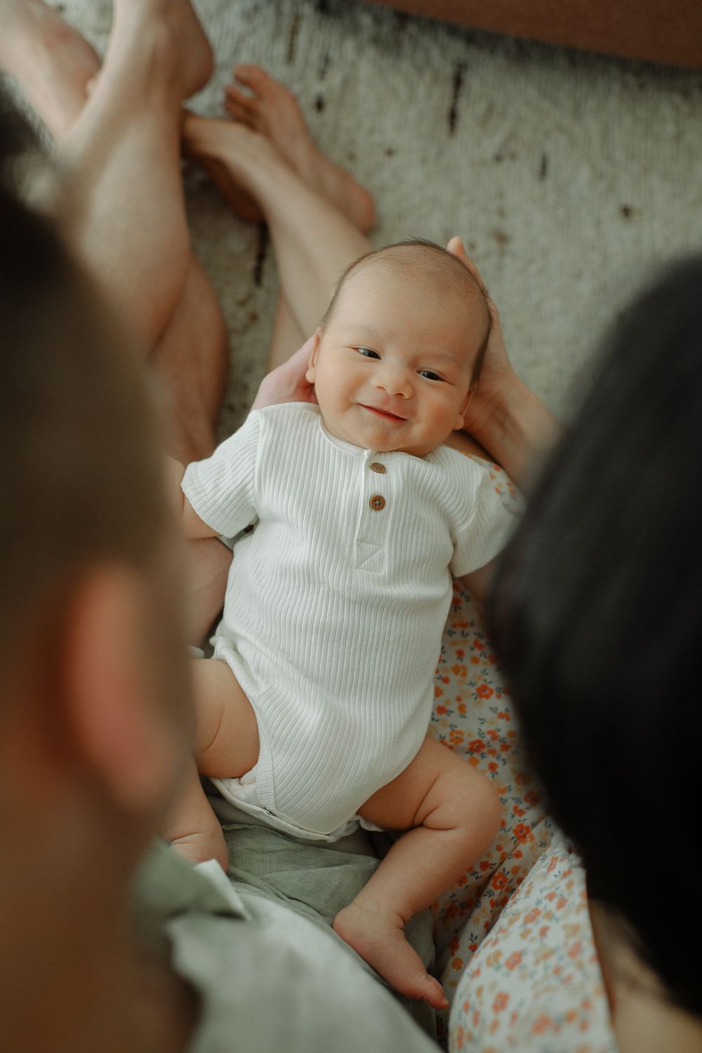 Baby wearing white ribbed onesie smiles while lying on floral fabric in natural lighting.