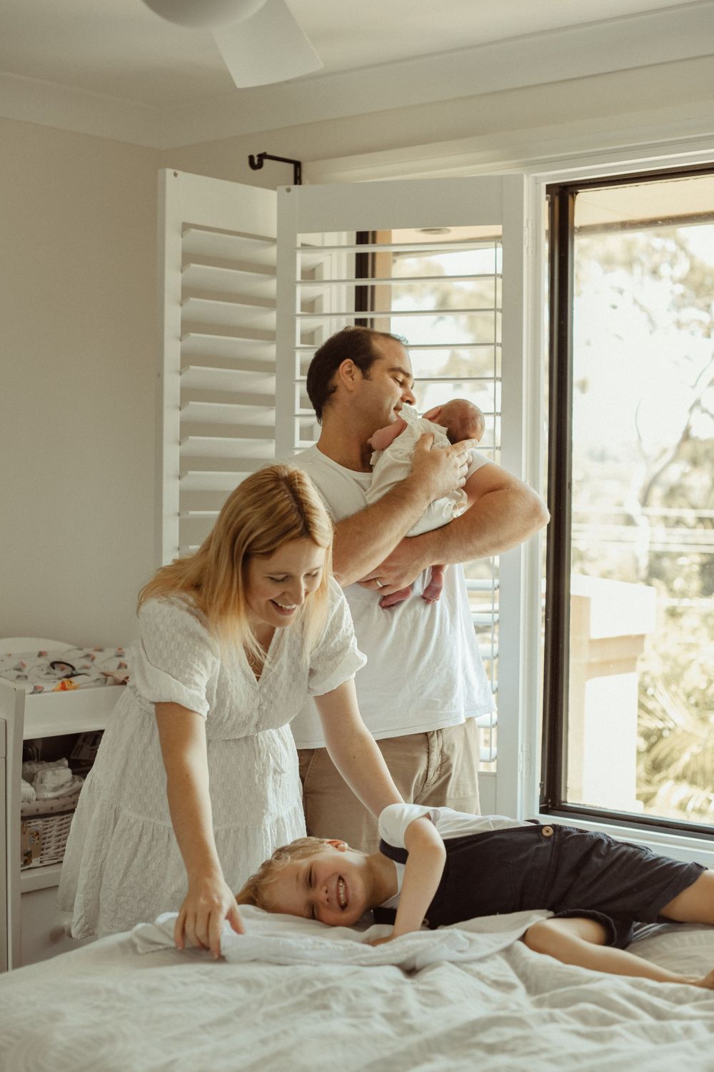 A joyful family scene on a bed with a toddler playing and a newborn being held.