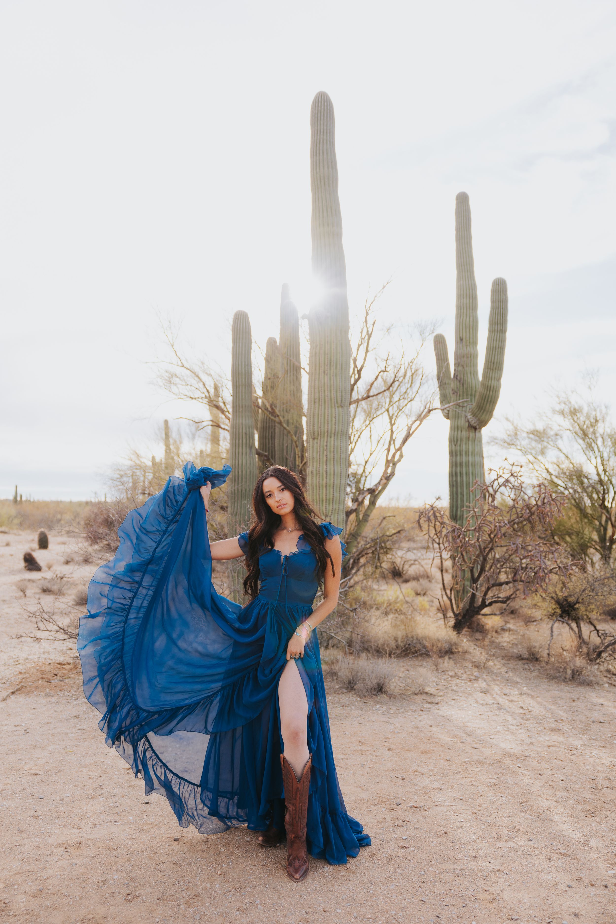 A flowing blue gown creates dramatic movement against the desert landscape with tall saguaro cacti.