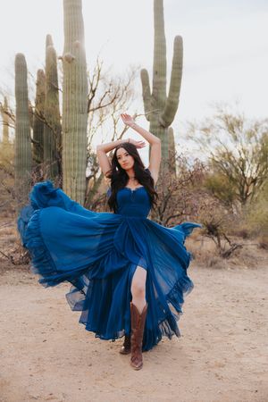 A dramatic blue gown floats gracefully in a desert setting with towering saguaro cacti.
