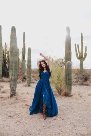 Elegant poses in a flowing blue dress contrast against desert cacti and sandy terrain.