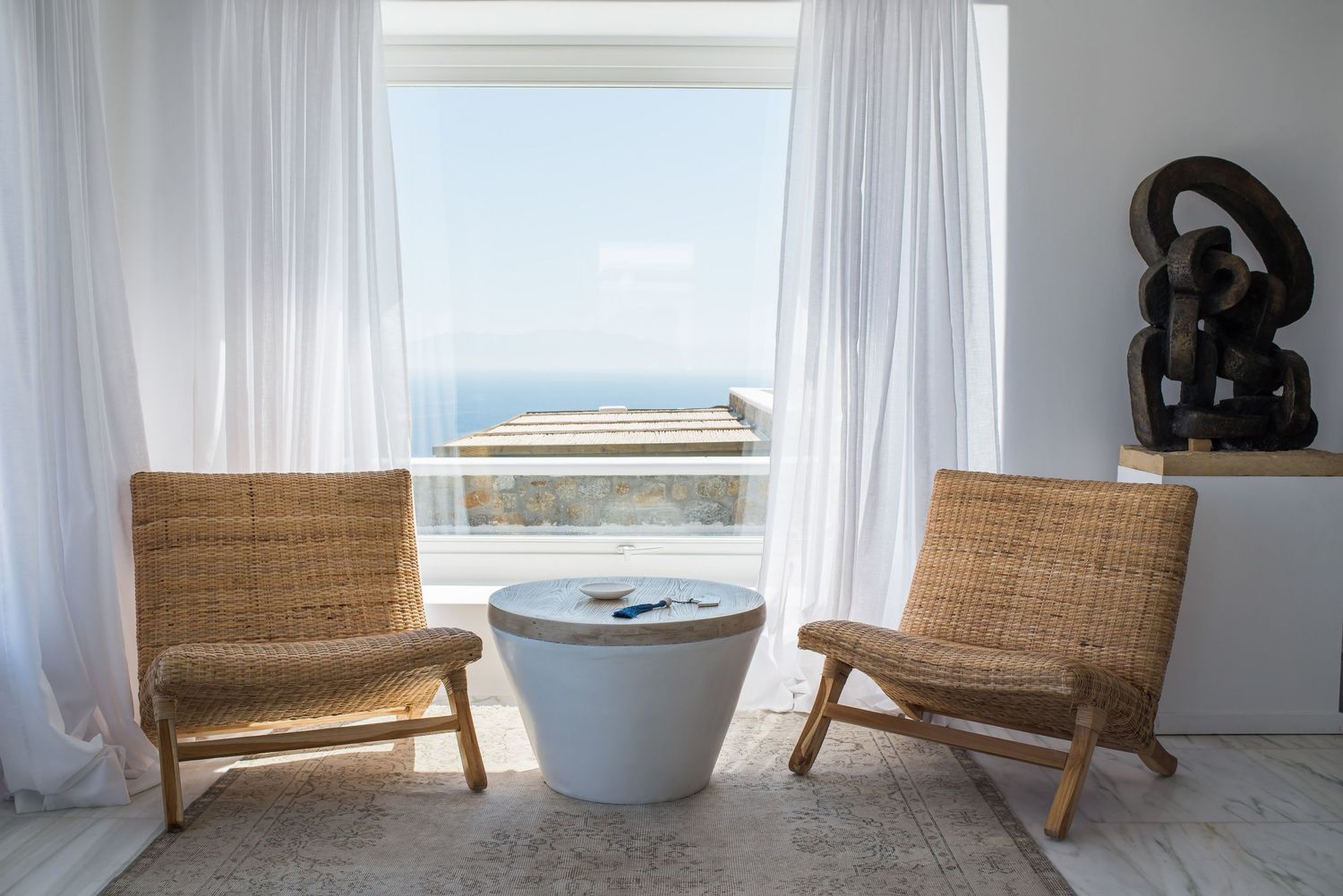 Two woven rattan chairs flank a white side table by a window with sheer curtains overlooking an ocean view. Villa in Mykonos