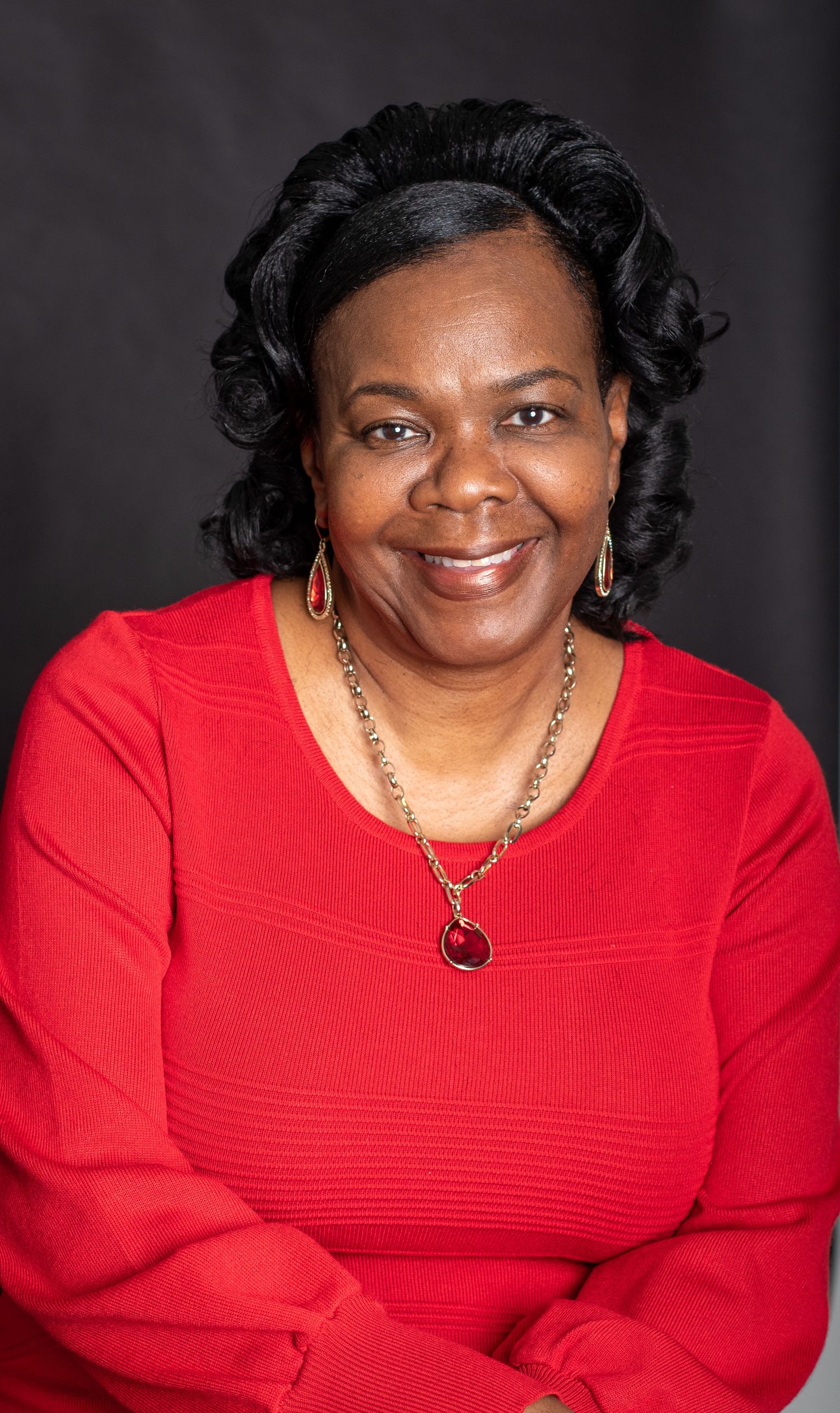Professional portrait of a person in a red sweater with curly black hair and jewelry smiling warmly against dark background.