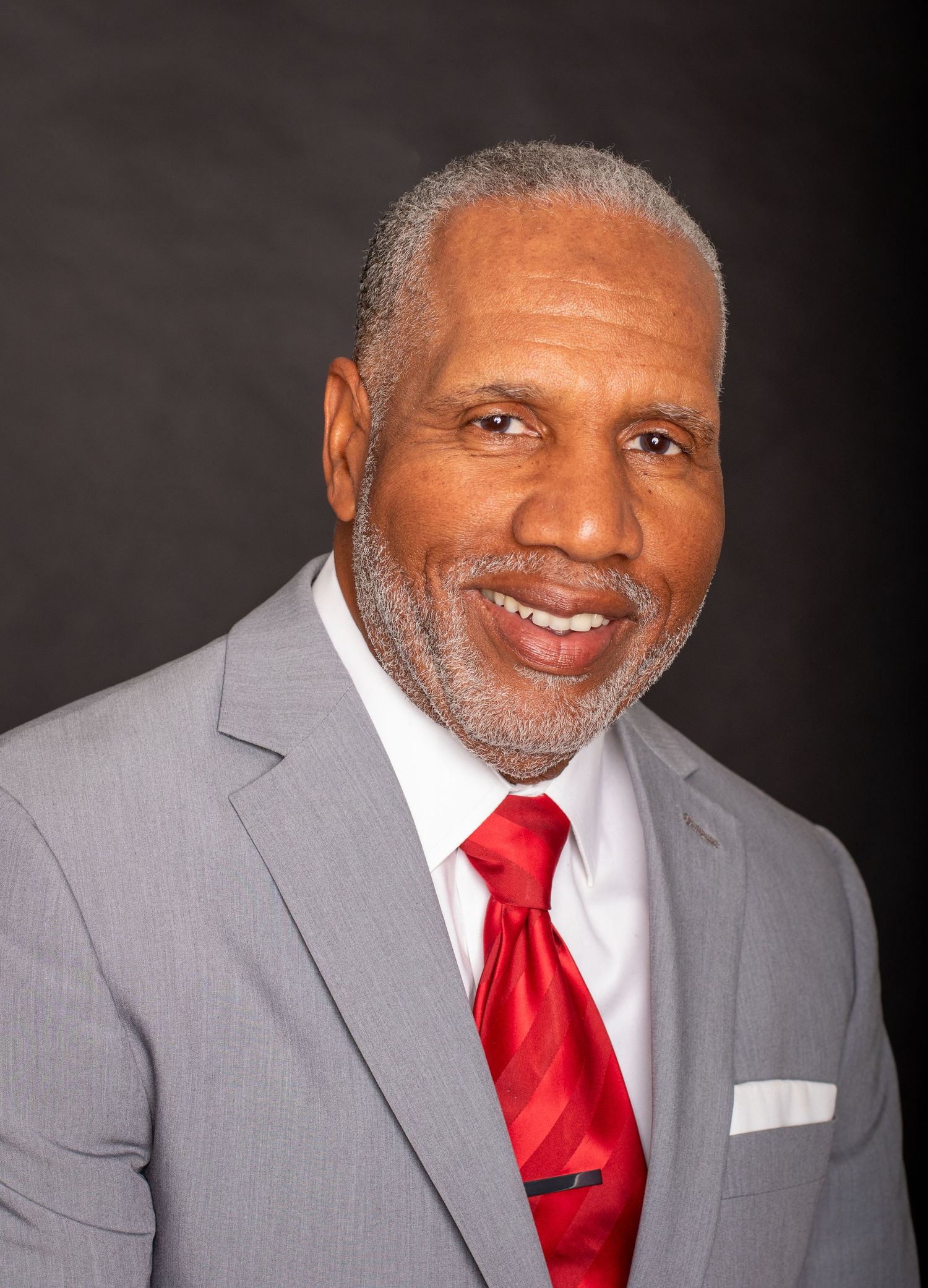 Professional headshot of person in grey suit with red tie smiling warmly against dark background.