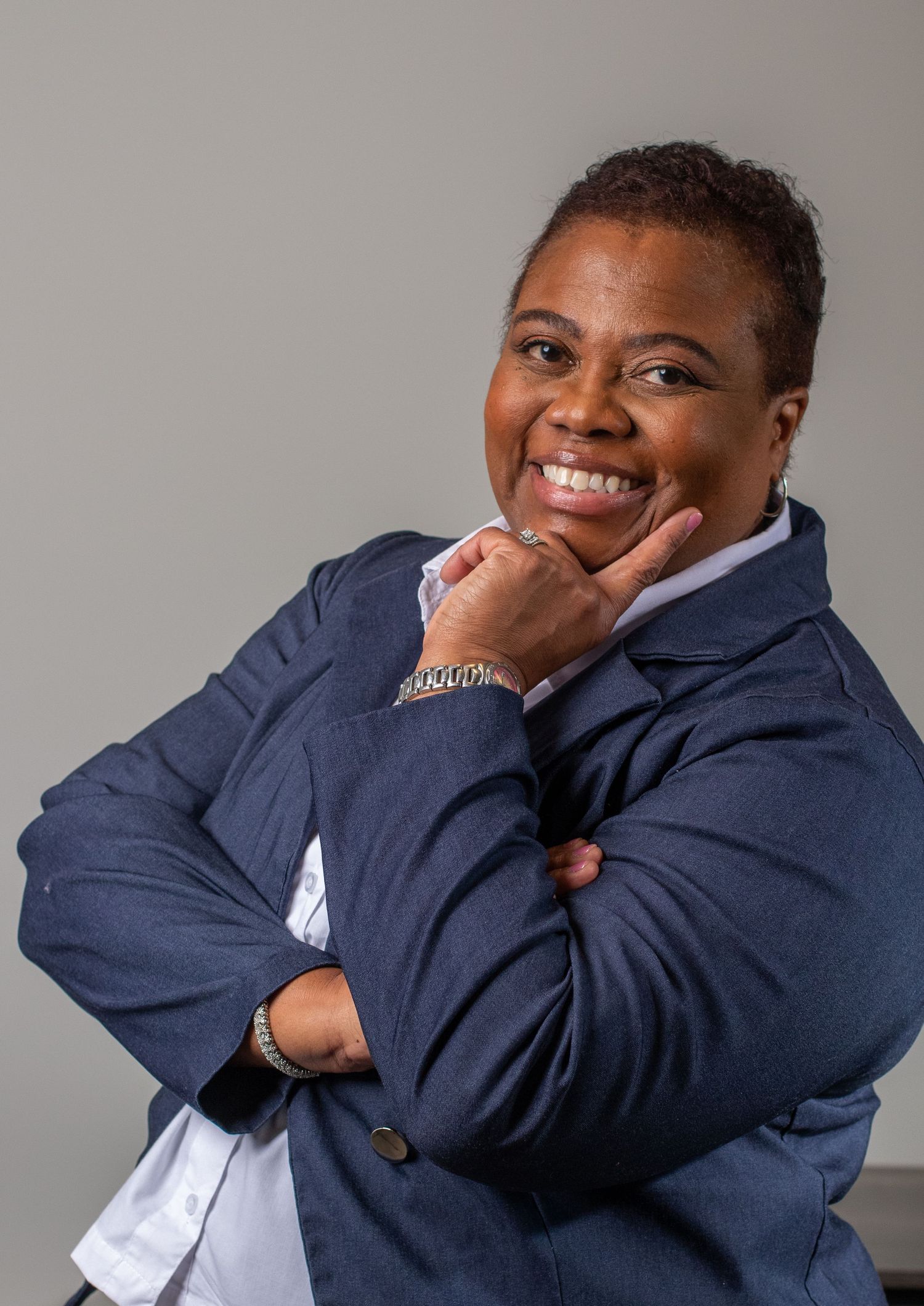 Person in navy blazer poses with hand under chin against grey background.