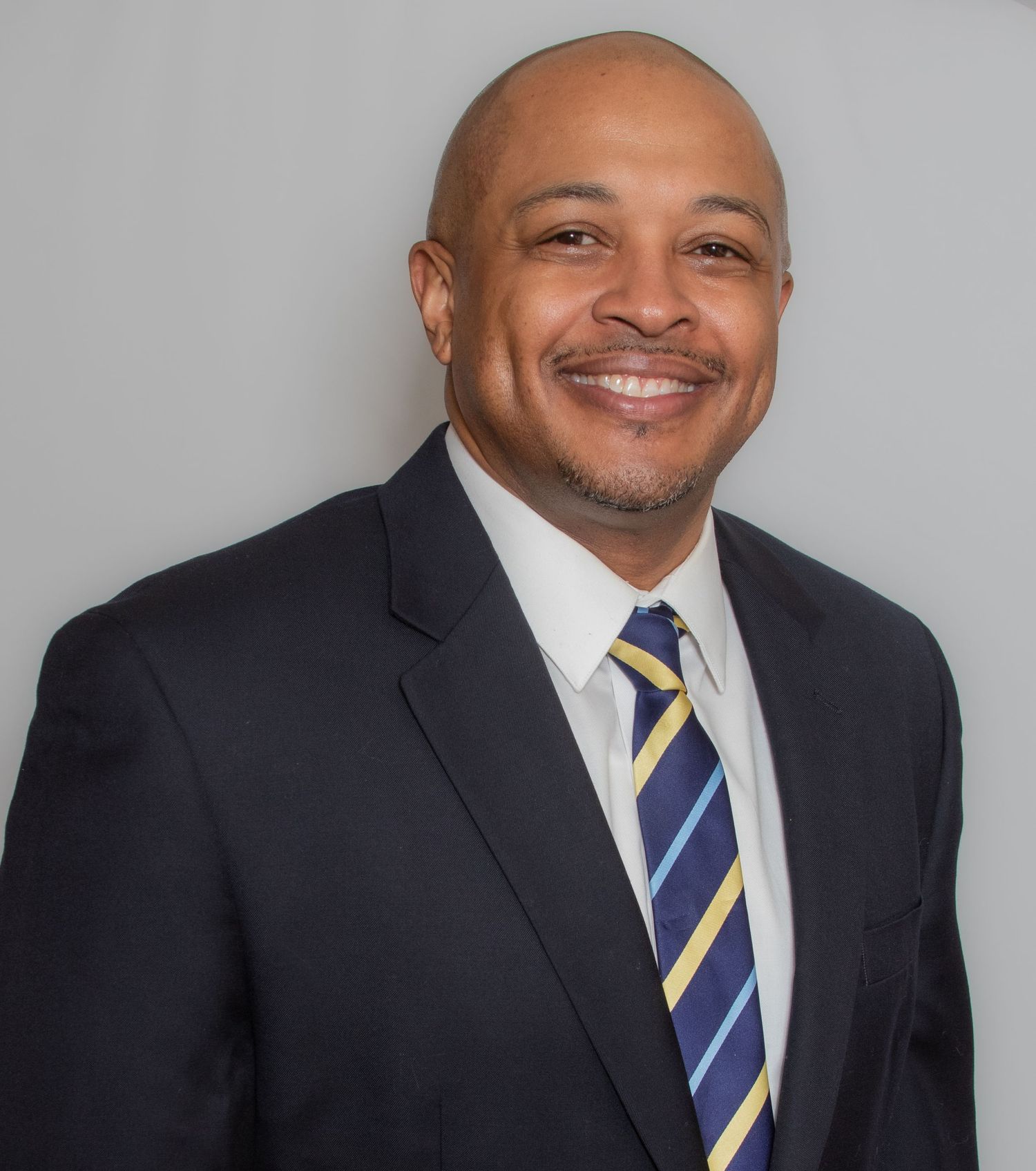 Professional portrait of person in dark suit with striped tie smiling against grey background.