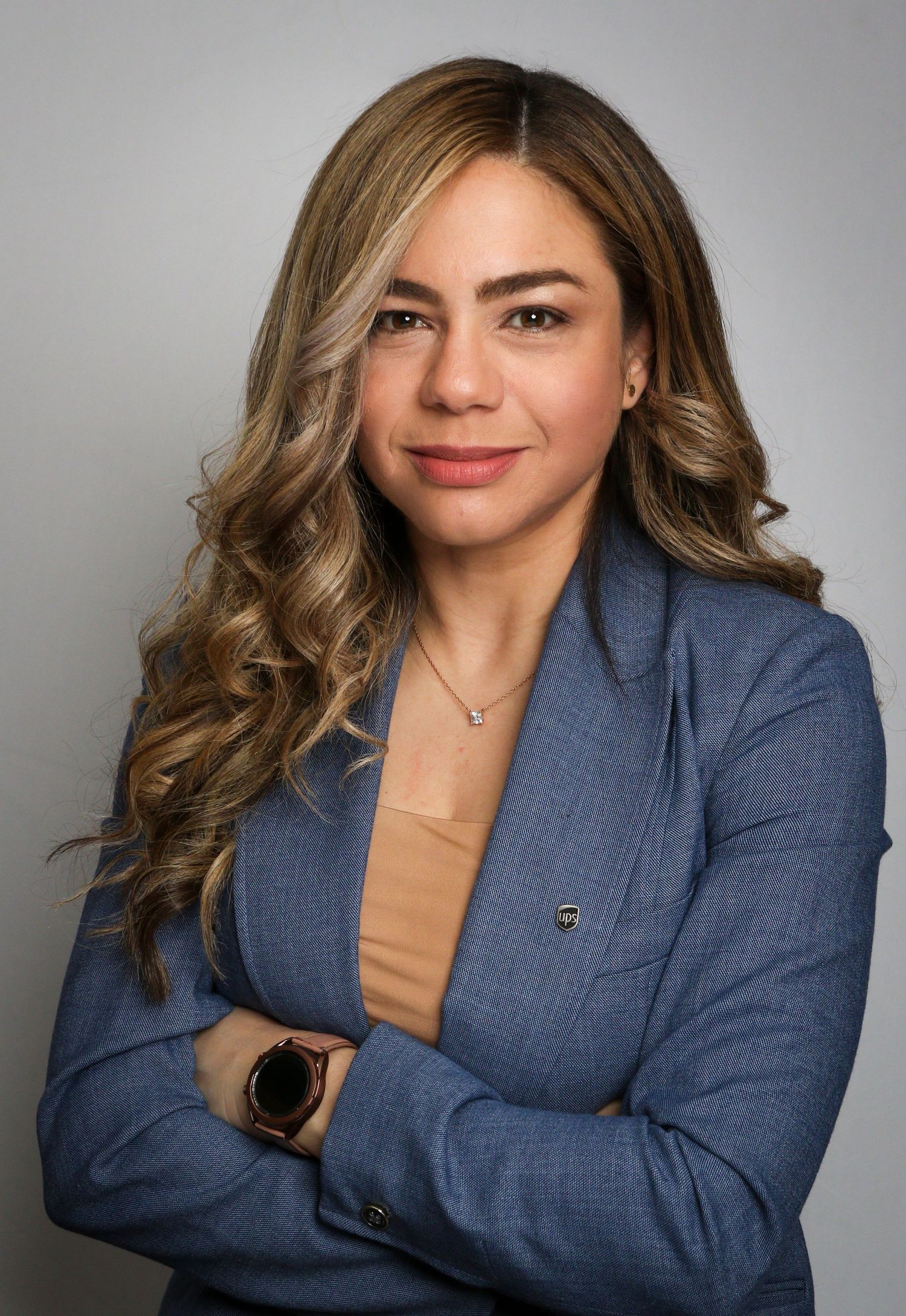 Professional headshot of person in blue blazer and tan top with styled wavy hair against grey background.