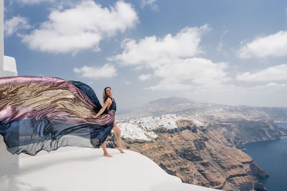 Flowing dress billows in the wind against stunning Santorini cliffs and blue Mediterranean waters.