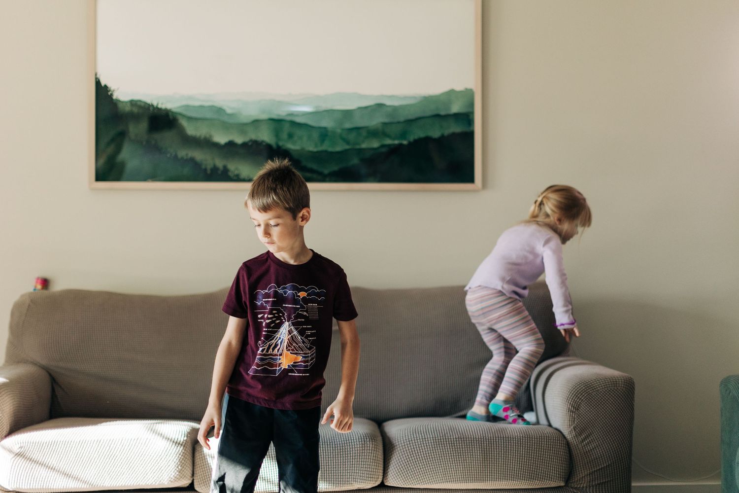 Two children playing energetically on a gray couch beneath a landscape painting.