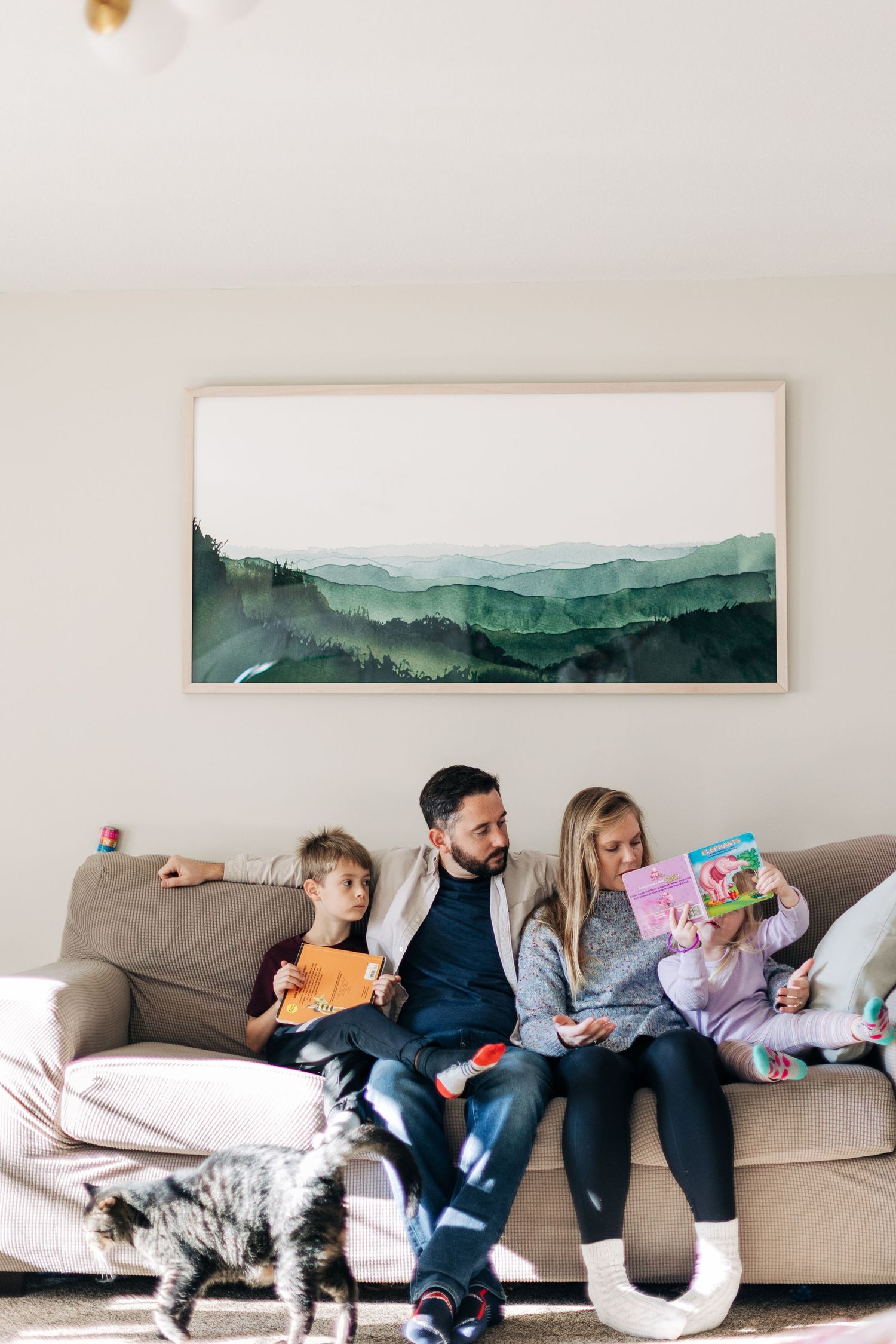 Family relaxing together on gray couch beneath watercolor mountain artwork while cat walks by.