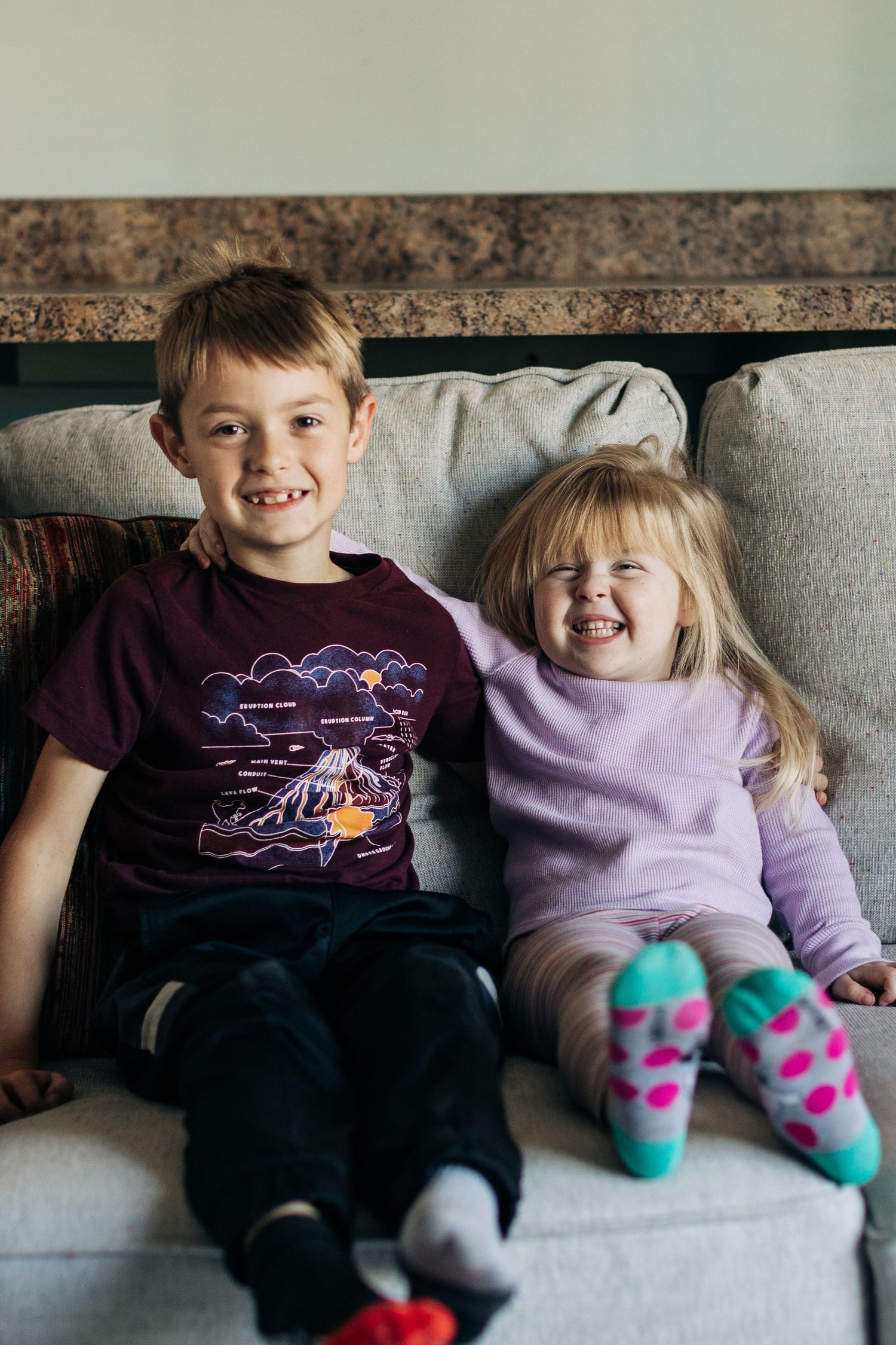 Two children sit side by side on a grey couch, wearing casual clothes and bright socks.