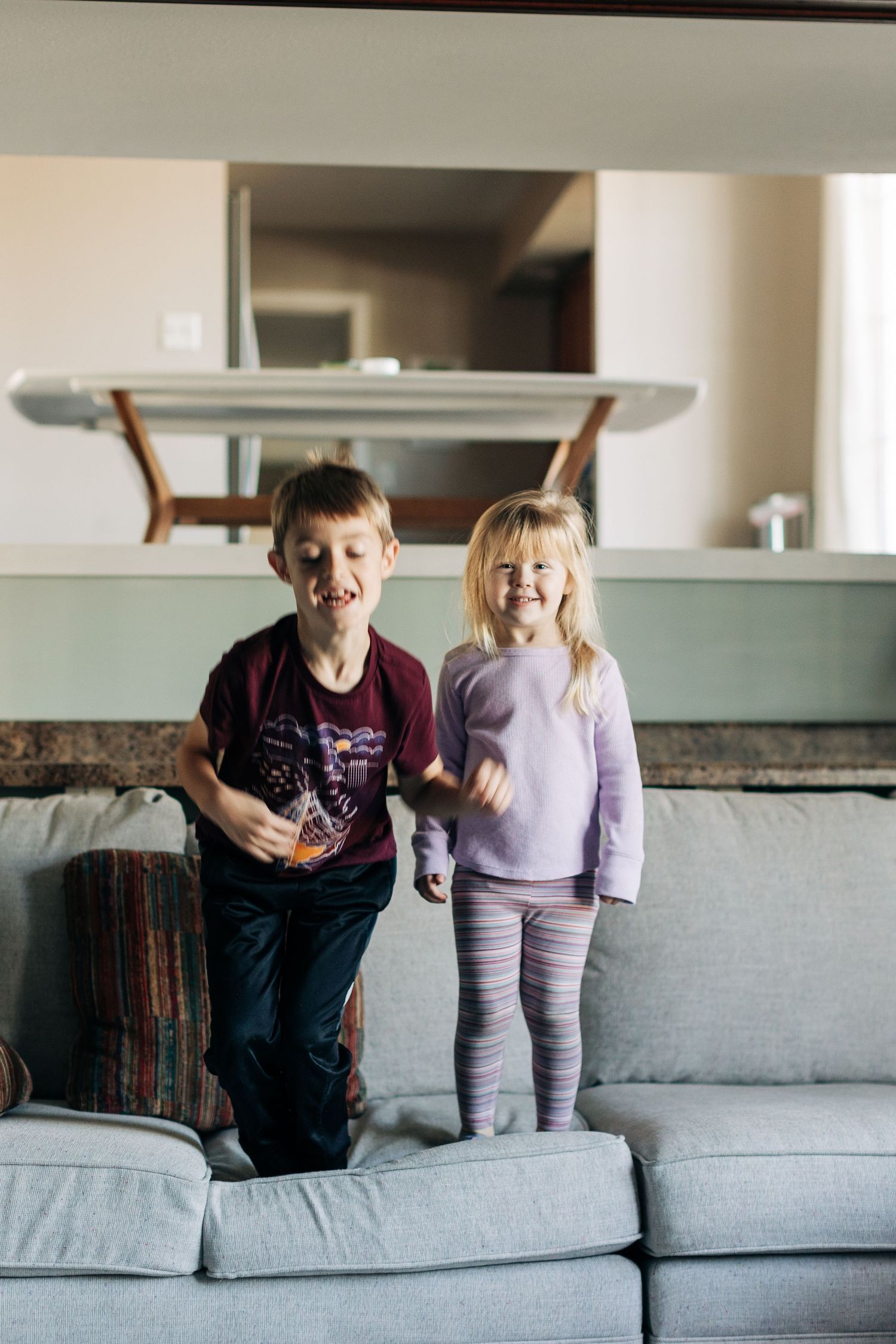 Two energetic kids standing on a grey sofa in a bright living room.