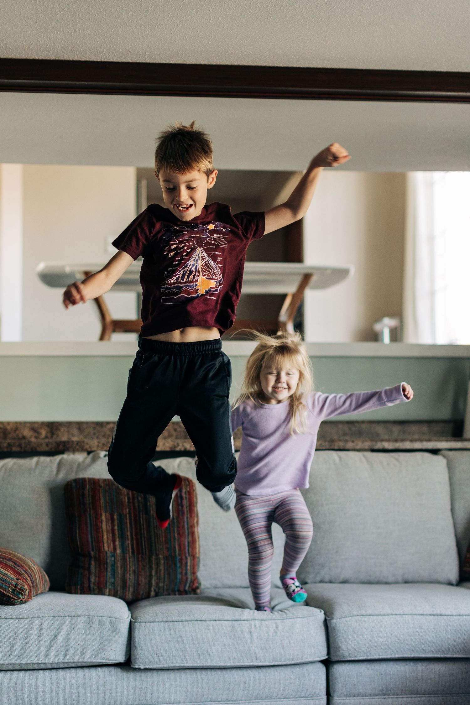 Kids jumping and playing on a grey couch in their living room.