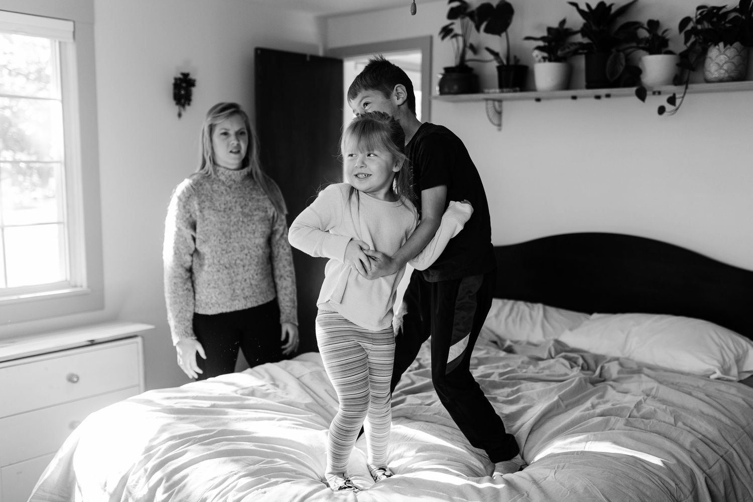 Family playtime on a white bed with indoor plants visible on wall shelving.