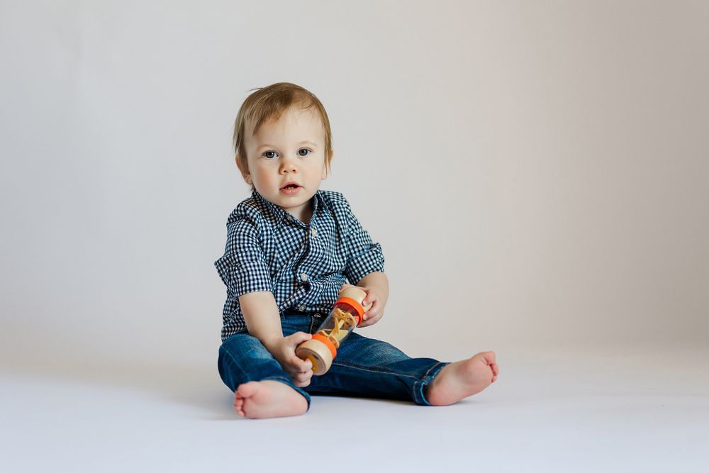 Baby in blue gingham shirt and jeans sits on white background playing with toy.