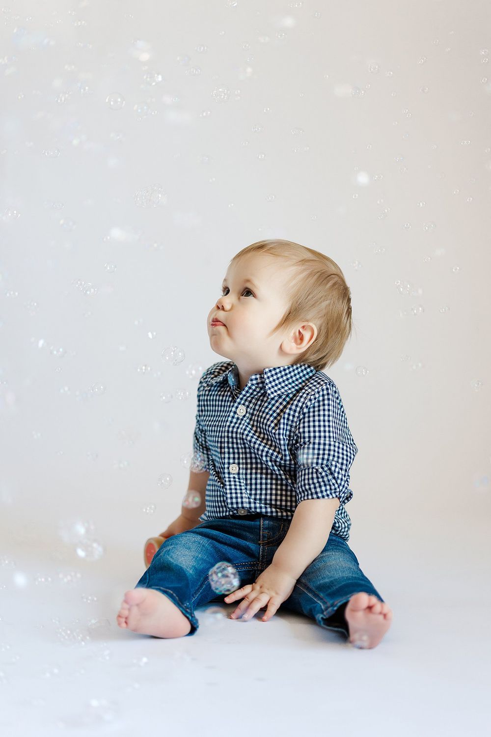 Baby in checkered shirt and denim crawls on white studio backdrop.
