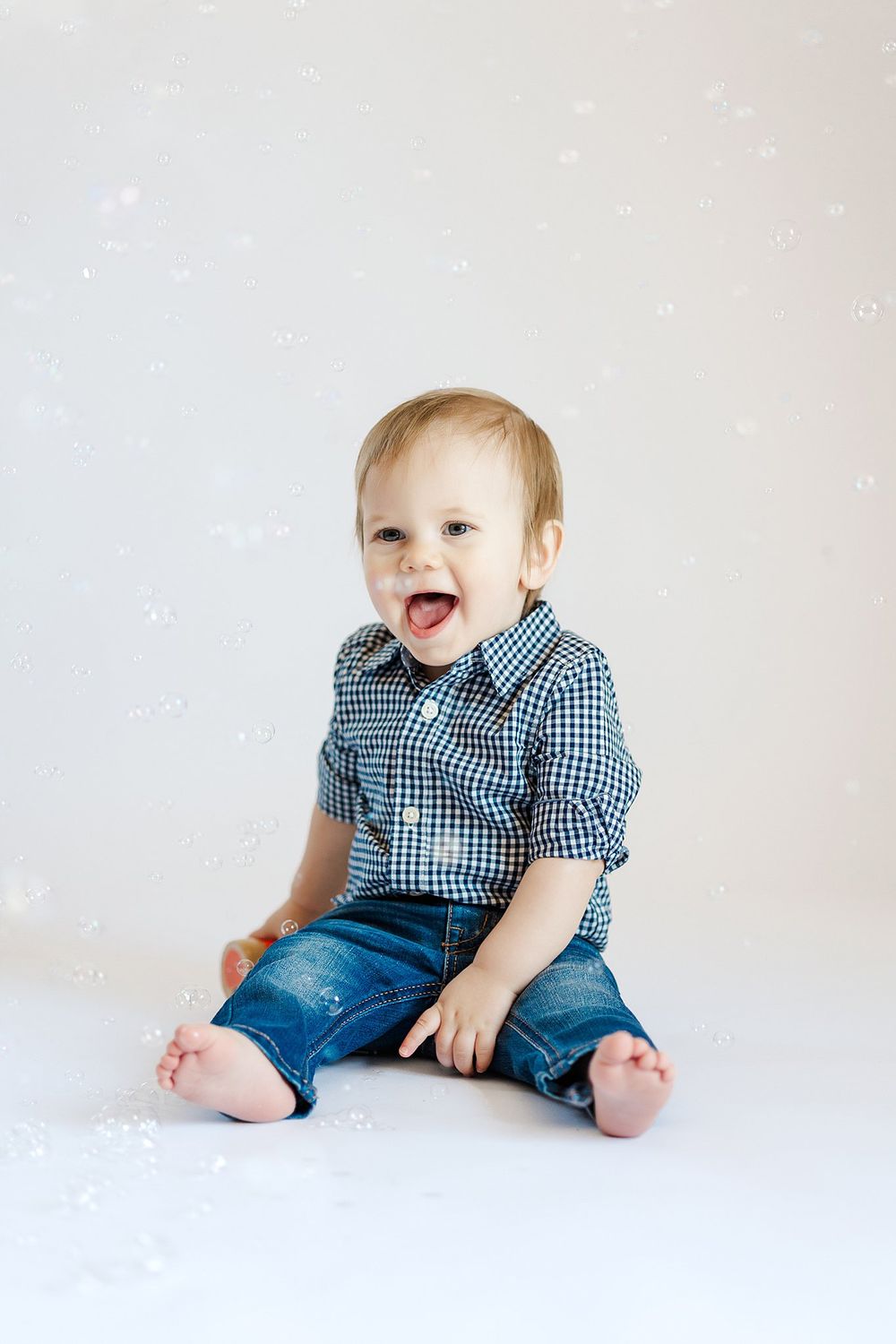 Laughing baby wearing blue gingham shirt and jeans sits on white floor.