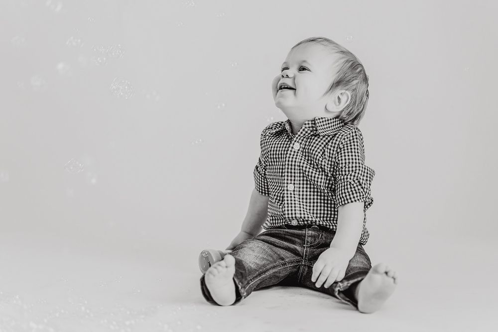 Baby in blue checkered outfit sits and plays on light background.