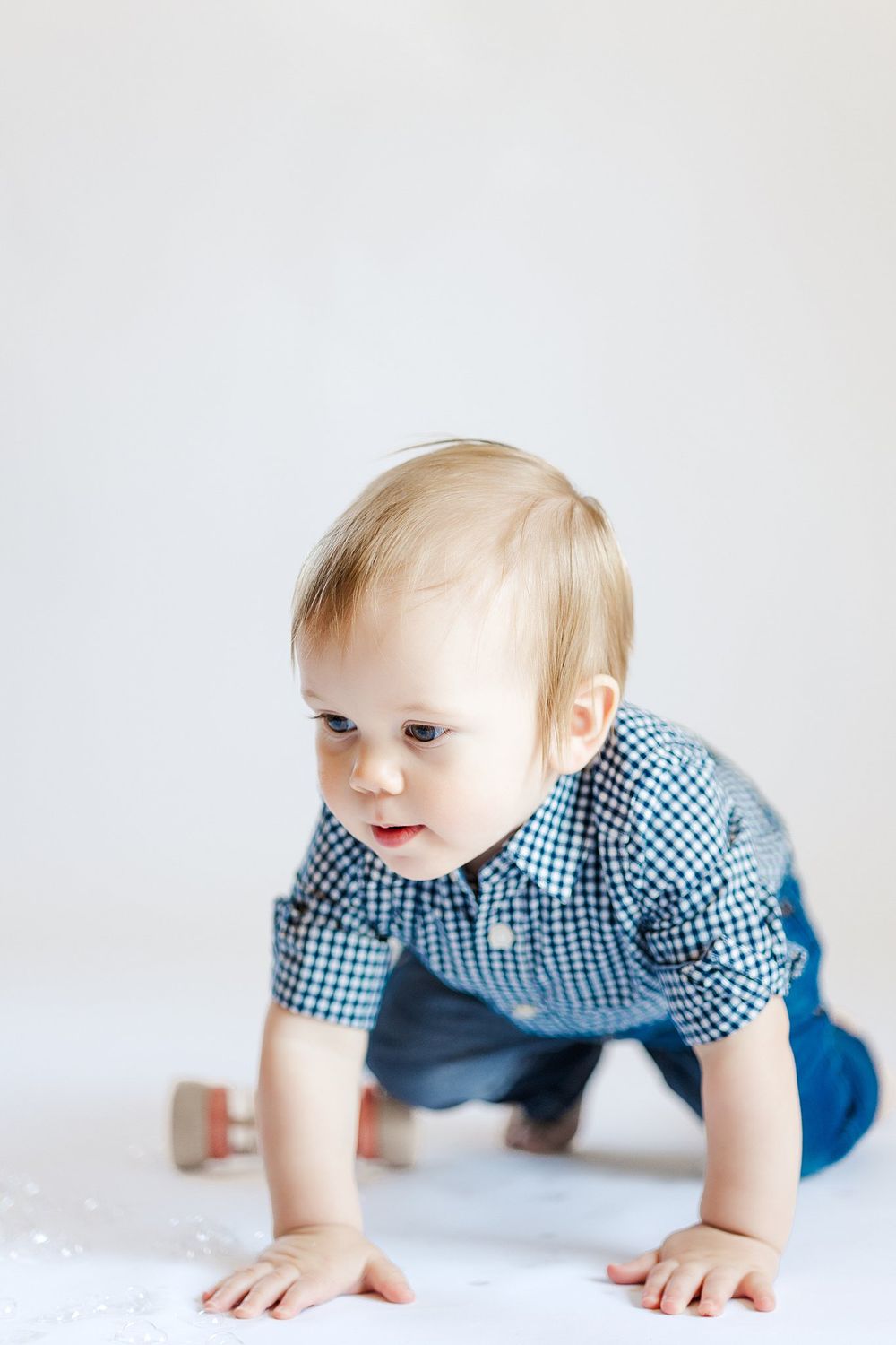 Baby in gingham shirt crawls on white studio floor during photoshoot.