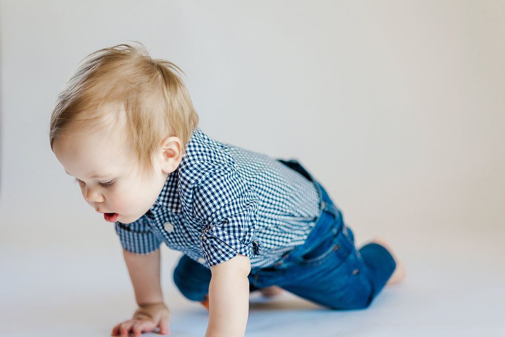 Baby in blue outfit crawls towards toy on white studio floor.