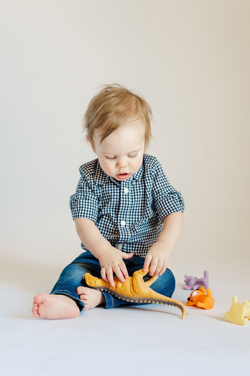 Baby in blue gingham shirt plays with toy dinosaurs on white background.