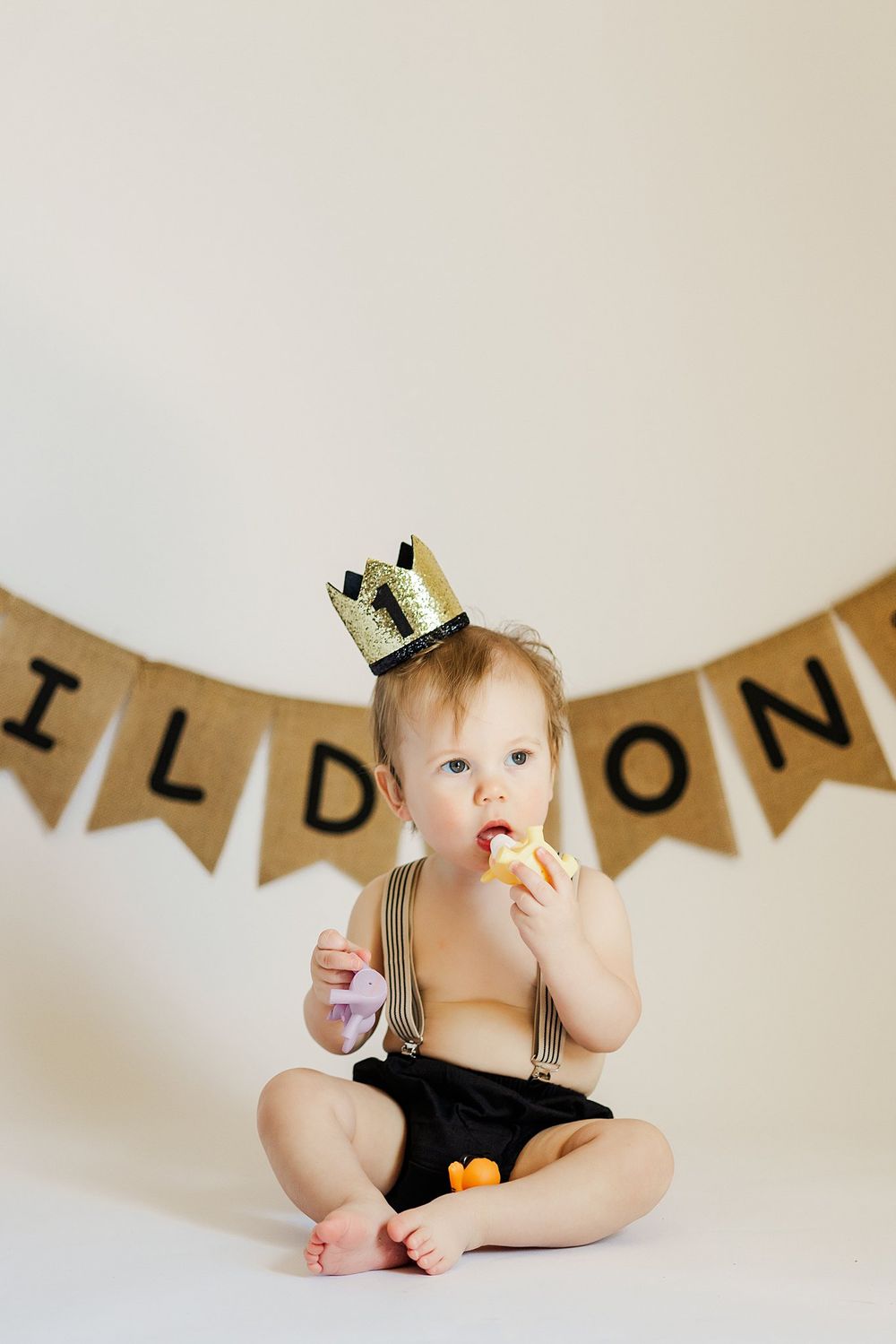 Baby wearing crown and suspenders sitting under Wild One banner eating snack.