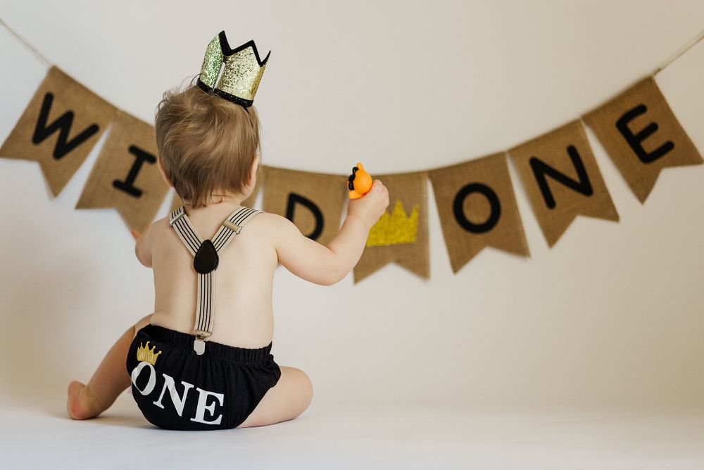 Baby in white outfit and crown reaching towards Wild One banner.