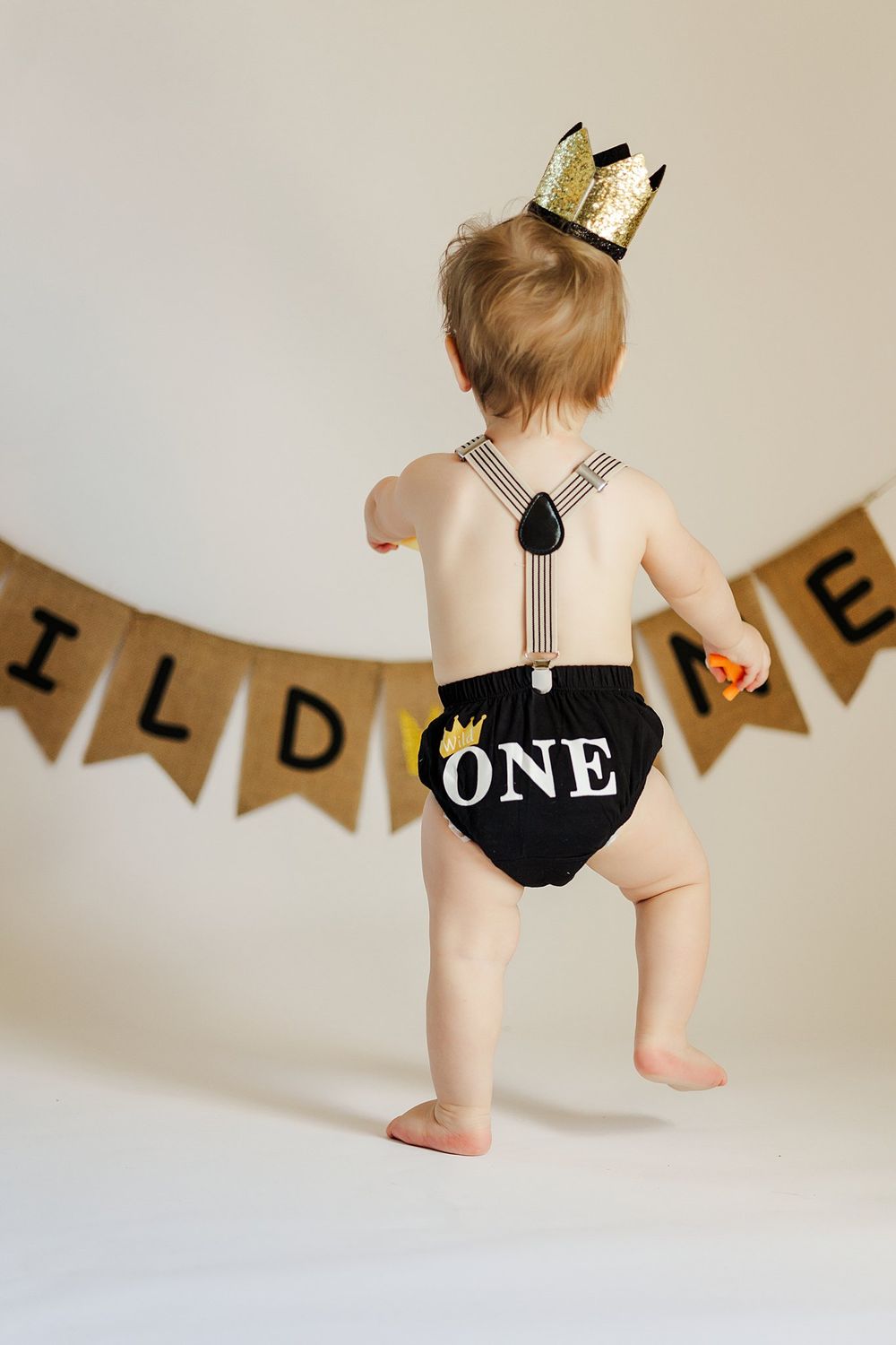 Baby in black diaper cover and gold crown standing in front of burlap banner.