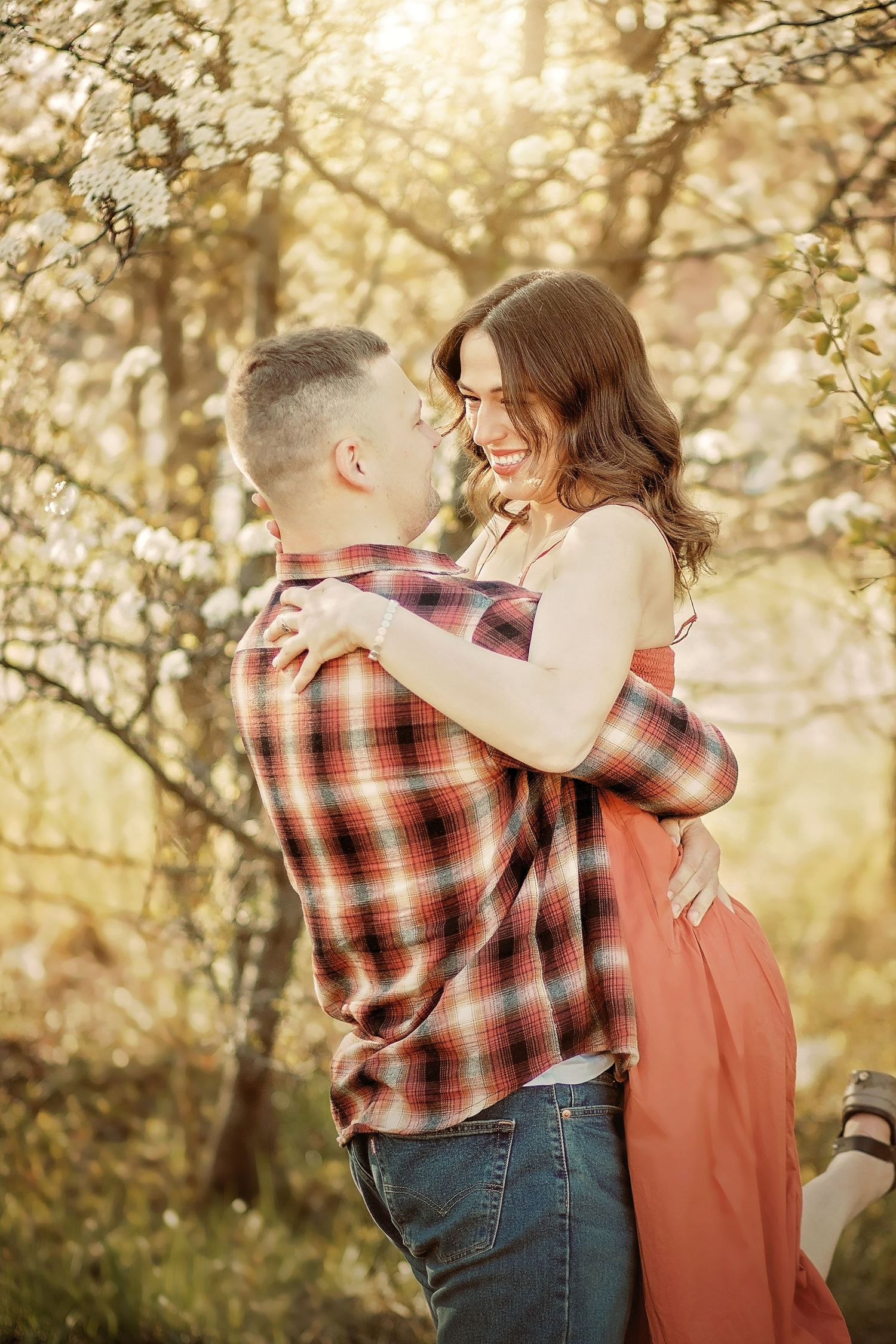 A smiling couple embraces tenderly among blooming spring trees, the woman in a coral dress and man in a plaid shirt. Spring Couple pictures