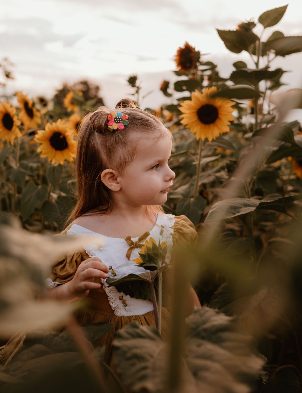 A young child explores a golden sunflower field at sunset while wearing a white dress and floral hair accessory.