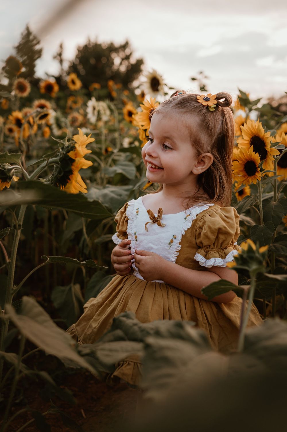A child wearing a ruffled dress smiles joyfully while standing among tall sunflowers at sunset.