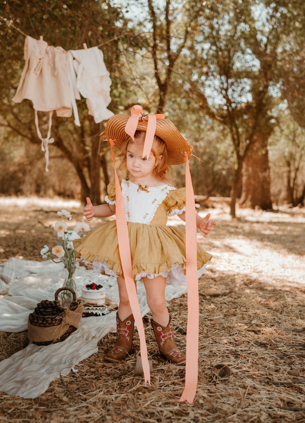 A child in a beige dress with pink suspenders plays outdoors in a rustic garden setting with warm lighting.