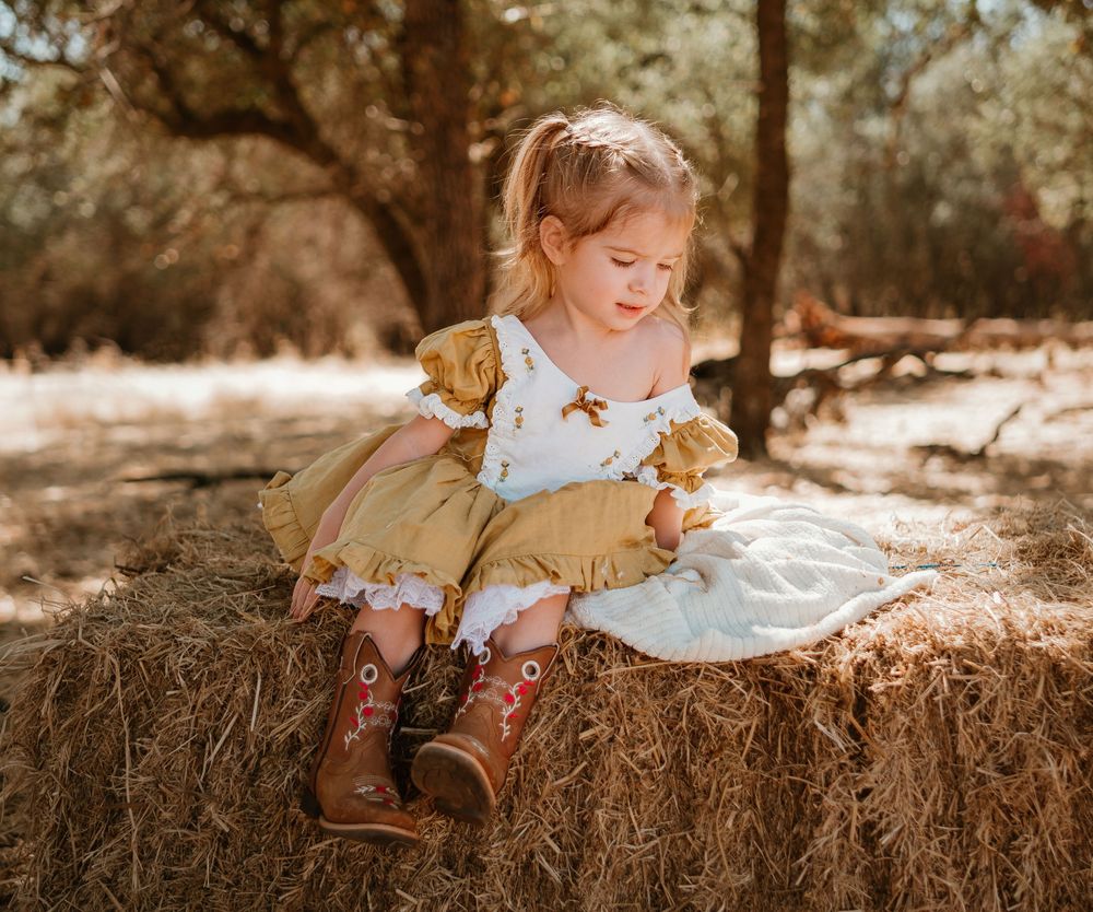 A child in a yellow dress sits on hay bales in warm sunlight during a rustic outdoor photo session.