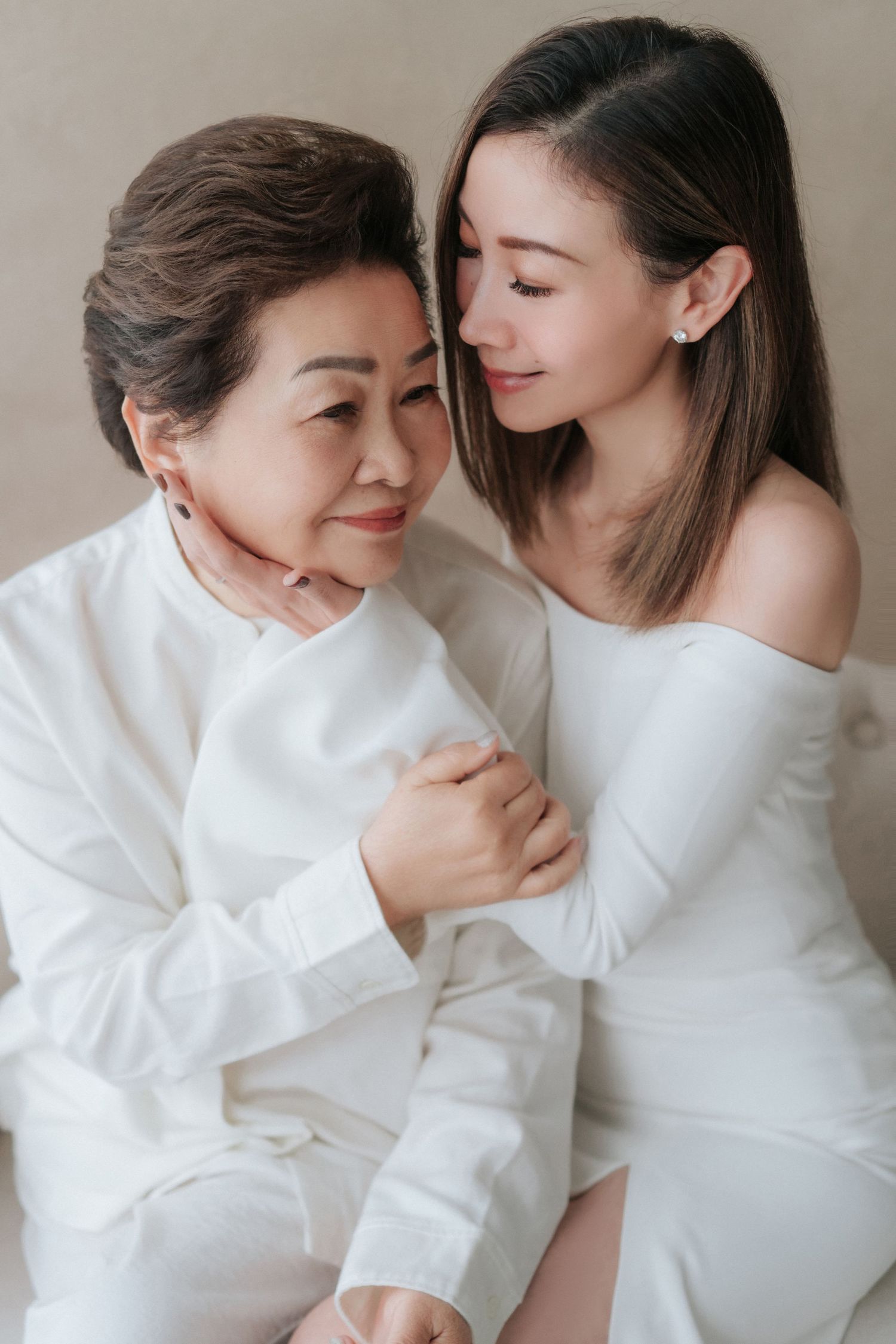 An intimate portrait of two women in white attire sharing a tender embrace against a neutral background.